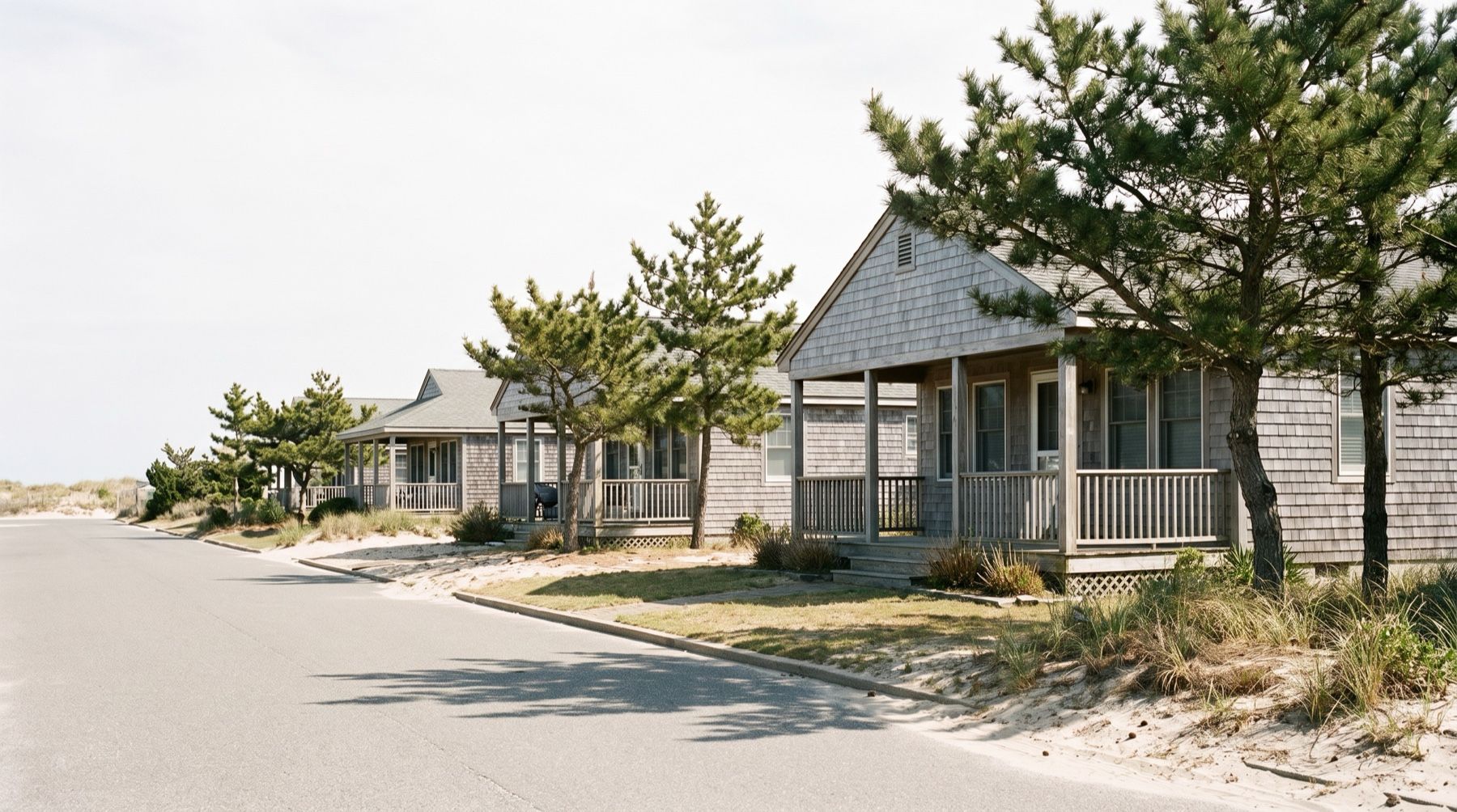 Low shingled cottages with covered porches behind sandy lawns and pine trees, under a bright overcast sky
